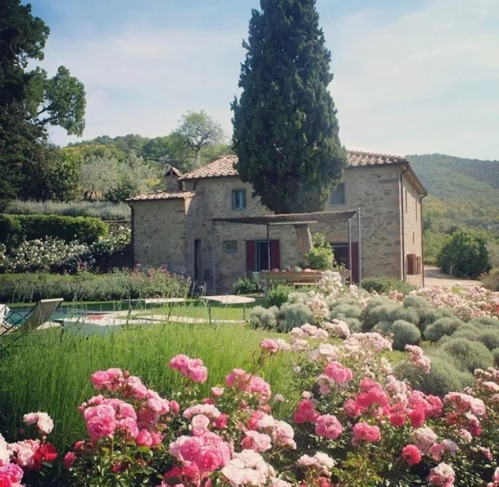 A rustic stone house with blooming flowers and tall trees in a serene countryside setting.