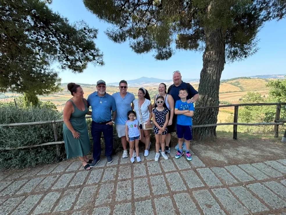A family poses together outdoors under two large trees with hills in the background.